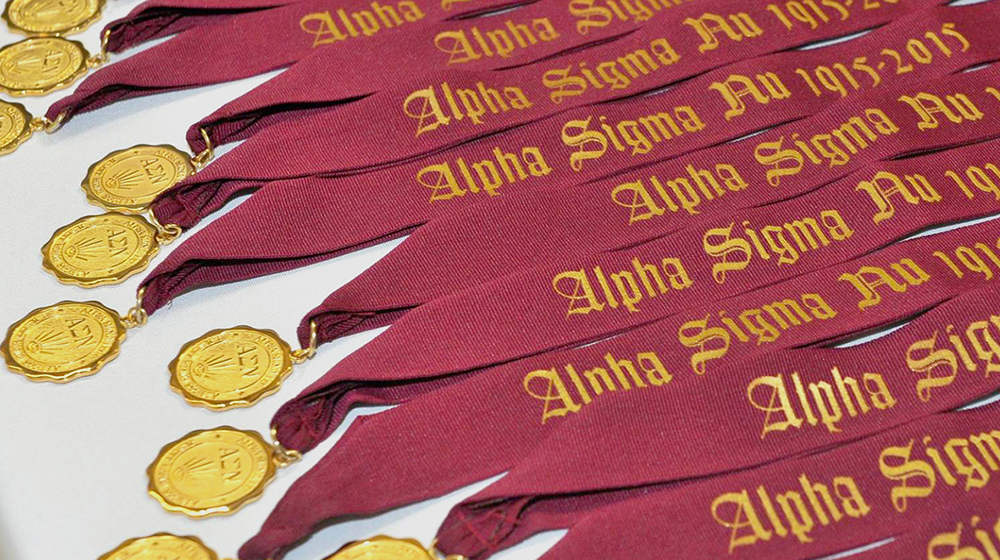 Alpha Sigma Nu honor society medals on maroon ribbons arranged in rows, with gold medallions displayed on a white surface