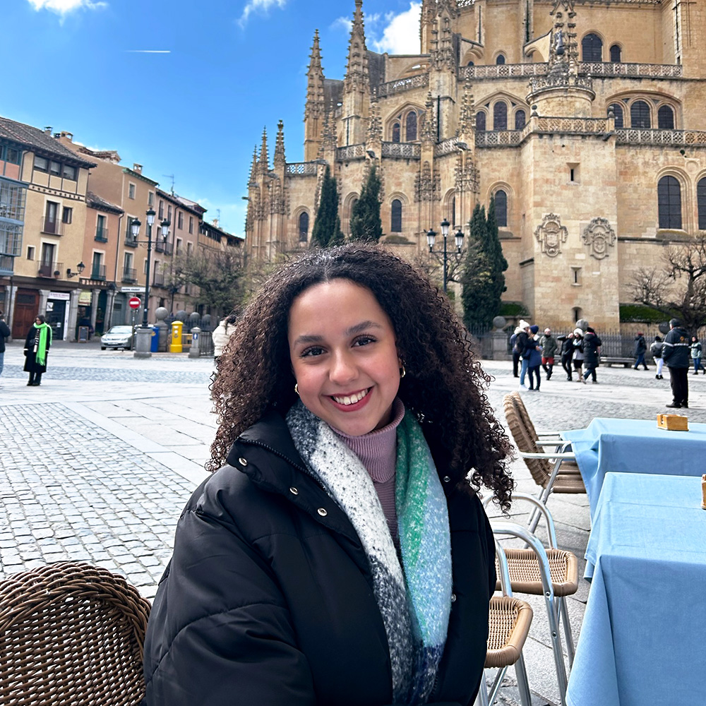 A smiling Becca Curiel sits at a table in a plaza, in Segovia, Spain, during her time studying abroad.