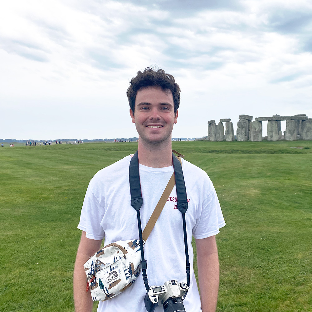 Ryan Maddigan, stands in an open grassy field in the UK. Stonehenge is in the background to his left.