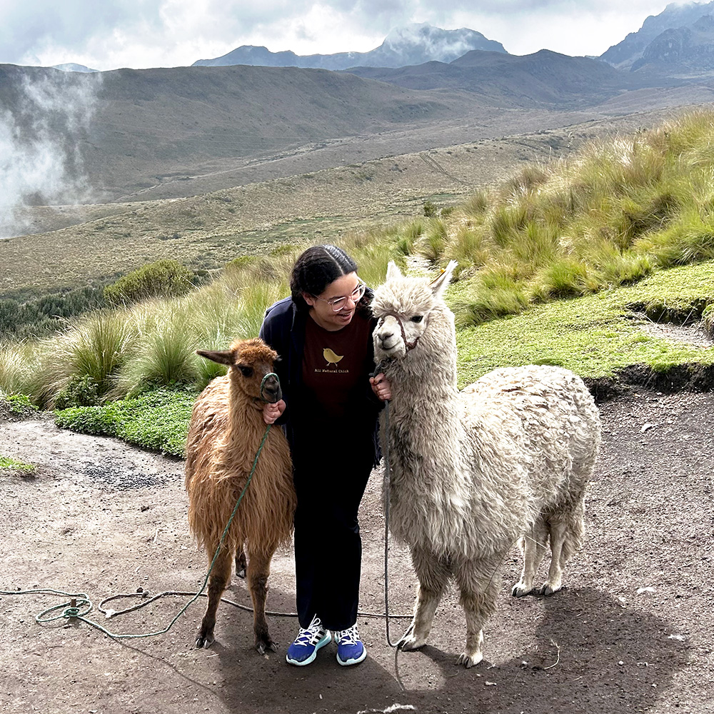 Salma Muhammad, in Quito, Ecuador stands with two llamas on either side of her. There is a mountain range in the background.