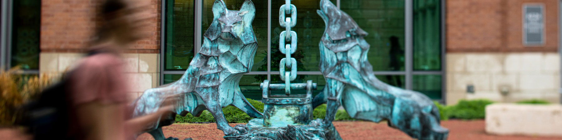 Bronze wolf statues with chain sculpture on campus, with a blurred student walking by
