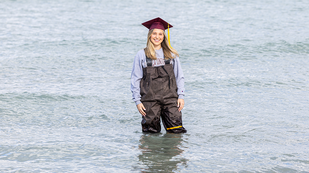 Mia Rogalski in waders in Lake Michigan wearing a graduation cap