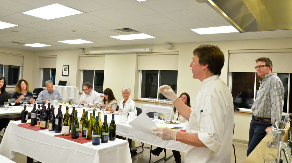 A chef leads a cooking class in a room and in the center is a table with multiple wine bottles