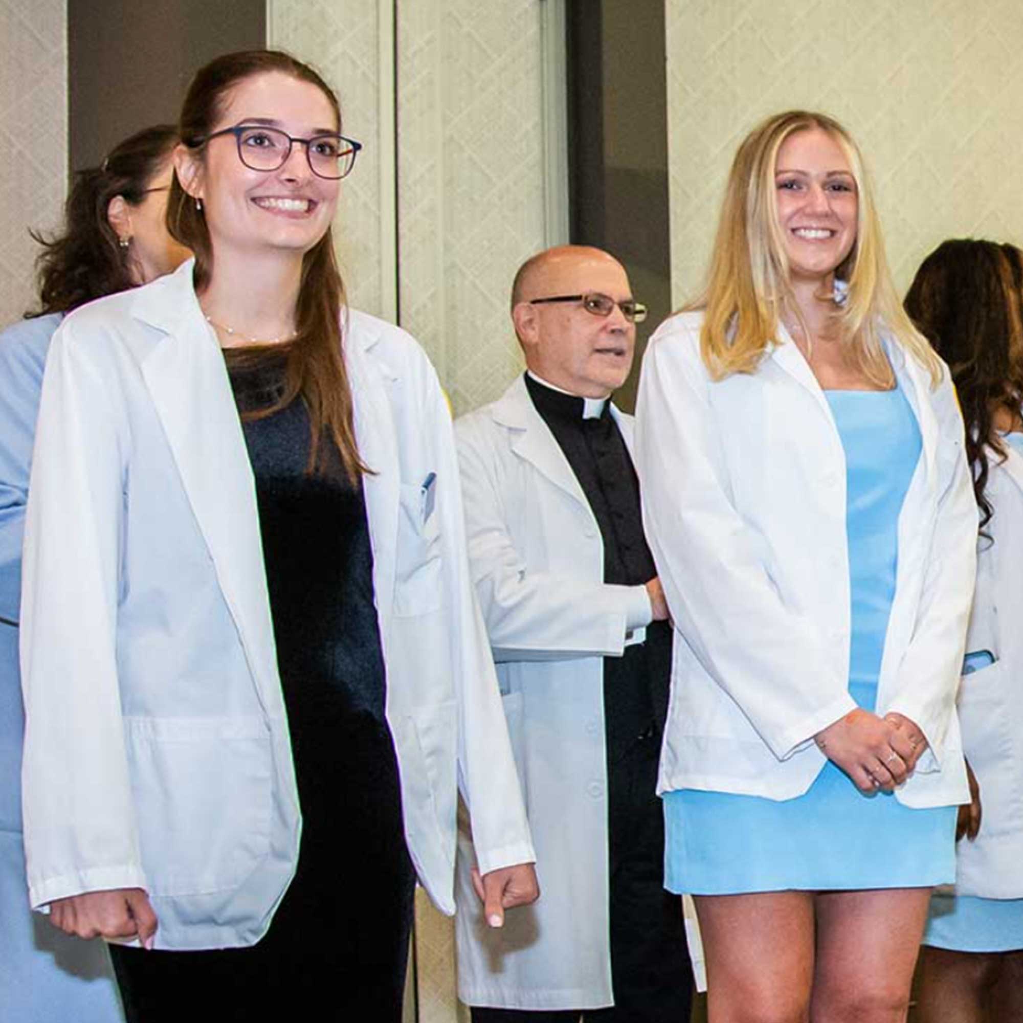 Two students wearing white coats with faculty members in white coats standing behind them